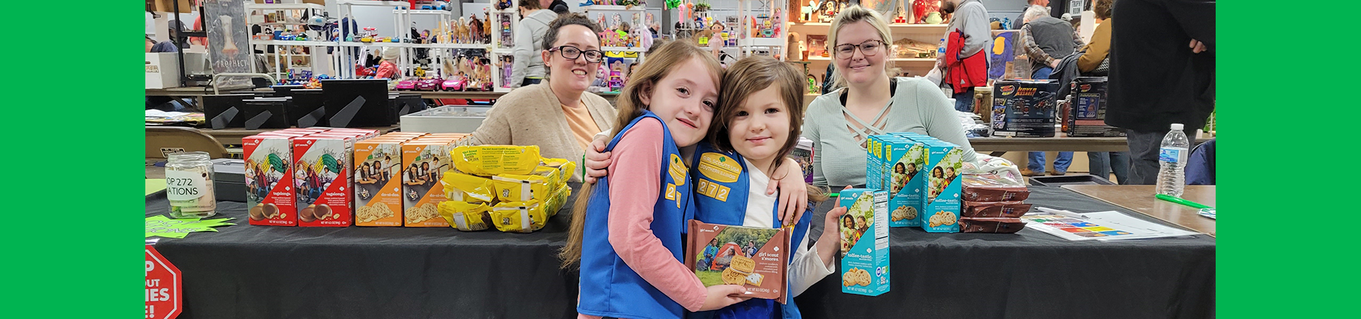  to girl scout daisies holding cookie boxes in front of a cookie booth with two adult volunteers sitting behind them 